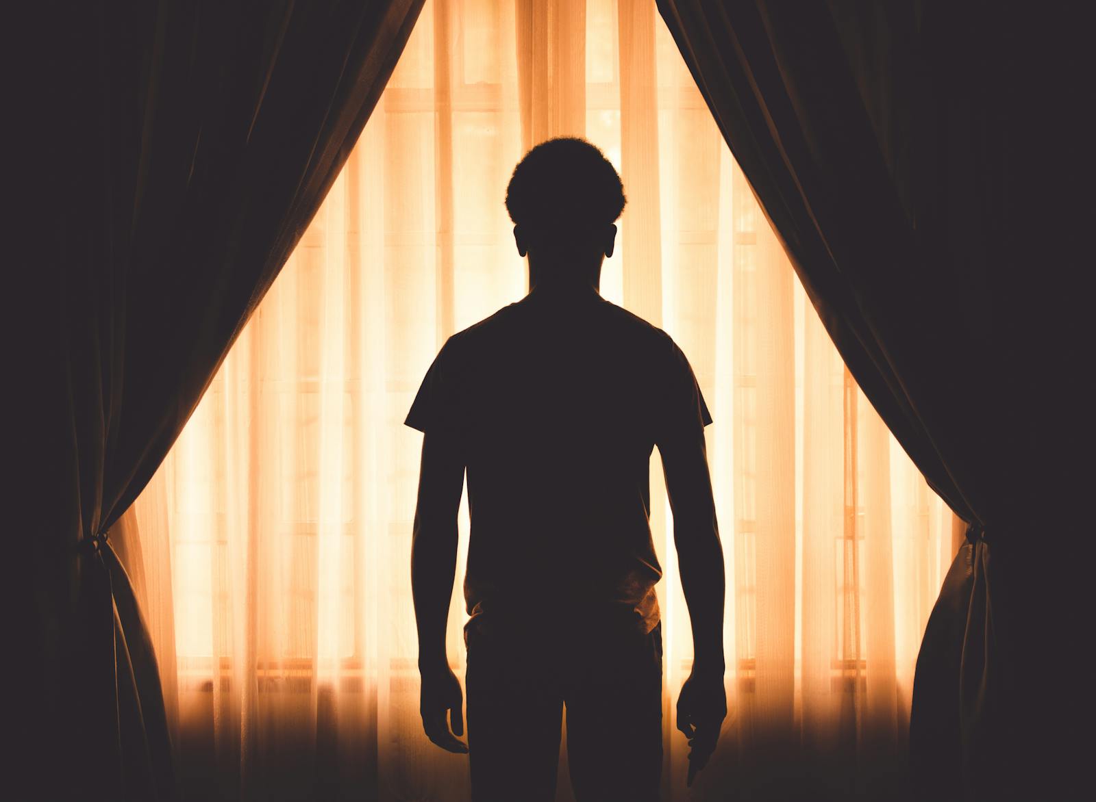A silhouette of a man standing by a window with curtains, creating a dramatic contrast and mood.