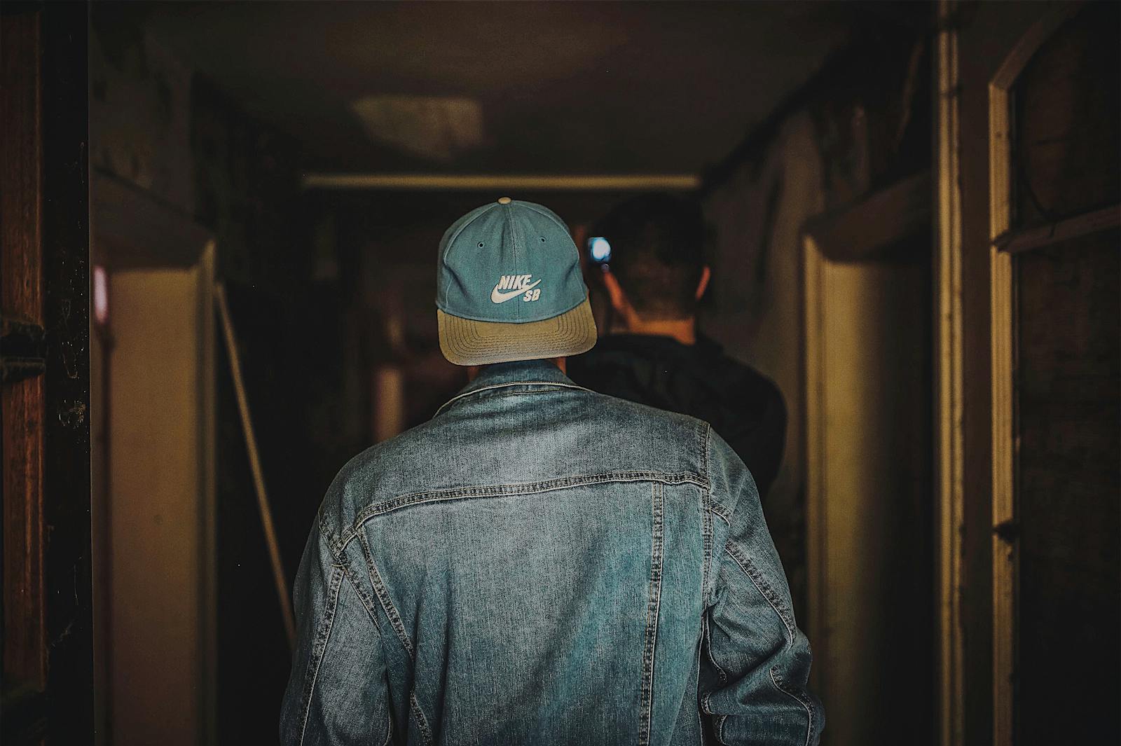 Back view of a man in a denim jacket and cap walking in a dimly lit hallway.