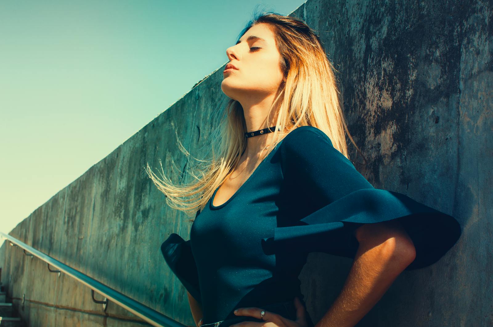 Fashionable woman posing with closed eyes against concrete wall, sunny day.