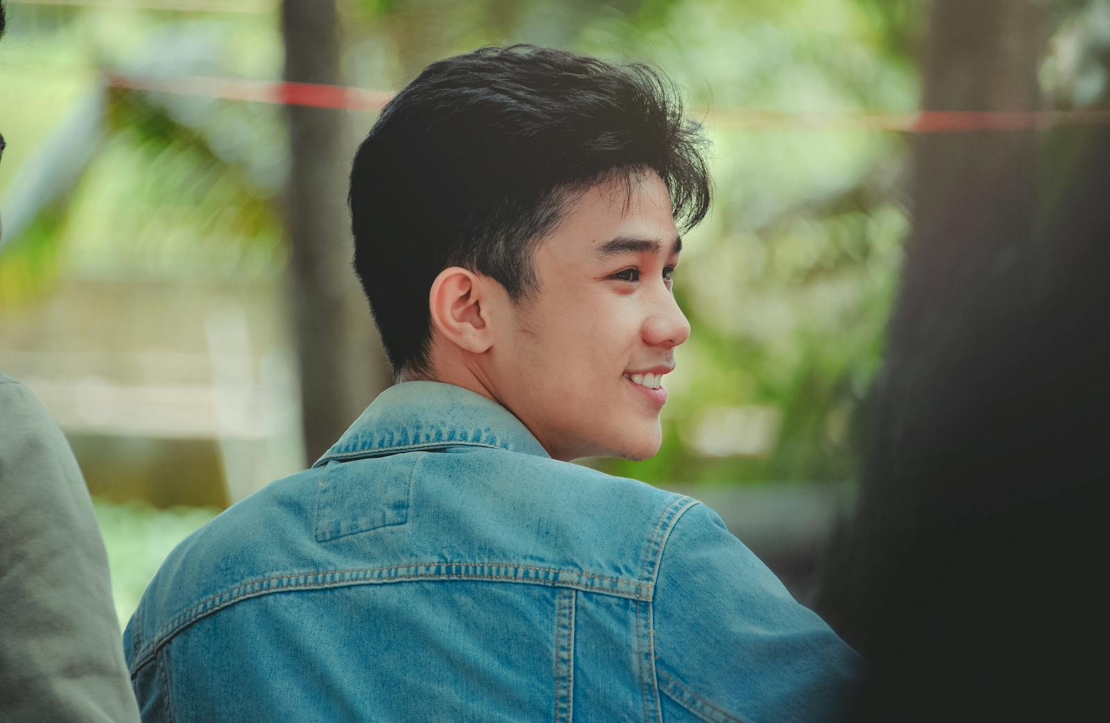 A young man with a joyful expression wearing a denim jacket in a natural setting.